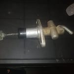 A metal automotive part, likely a clutch or brake master cylinder, featuring a cylindrical body, connecting rod, and attachment bracket, placed on a dark surface, possibly a workbench.