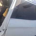A side view of a white vehicle, showing the rear door and window with tinted glass, partially revealing the interior which has a seat with torn upholstery. The background consists of grass and a house.