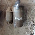 A close-up view of a used starter motor showing wear and dirt on its surface, with one side partly obscured, resting on a gritty floor.