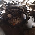A close-up view of a disassembled auto part, featuring a metallic gearbox with intricate mechanical details and components. The part is resting on a concrete floor, surrounded by other automotive parts.