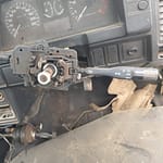 Close-up view of a car's steering column control, showing the steering wheel with gear and wiper control levers, part of a disassembled dashboard, and visible wiring beneath it.