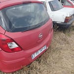 A red hatchback car with a dark rear window and a visible license plate parked in a grassy area, surrounded by other cars that appear abandoned.